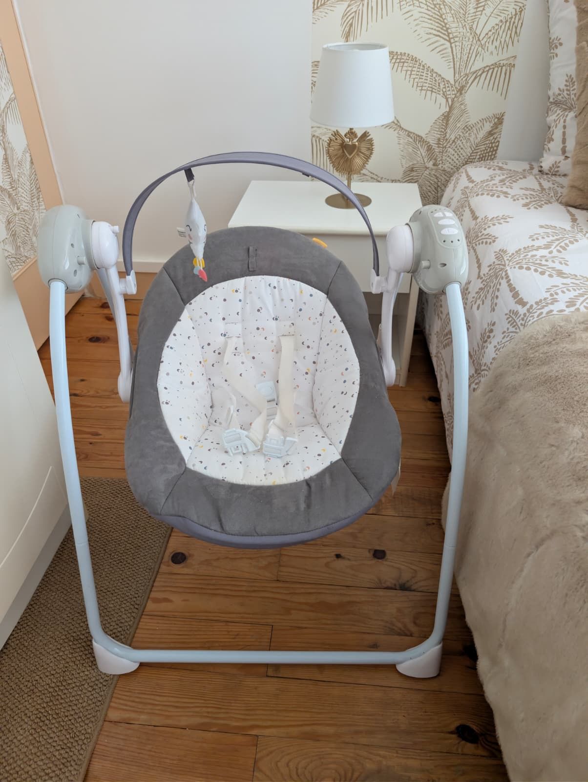 Un transat pour bébé avec une structure grise et un intérieur blanc parsemé de motifs colorés, posé sur un plancher en bois.
