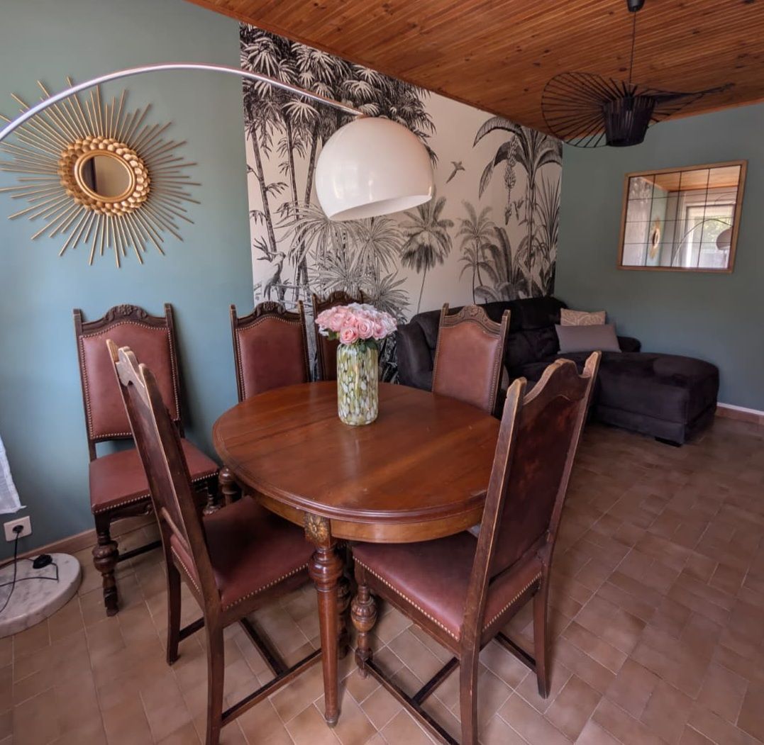 A dining area with a round wooden table, six chairs, a vase with flowers, a wall mural, a ceiling fan, and a window with a view.