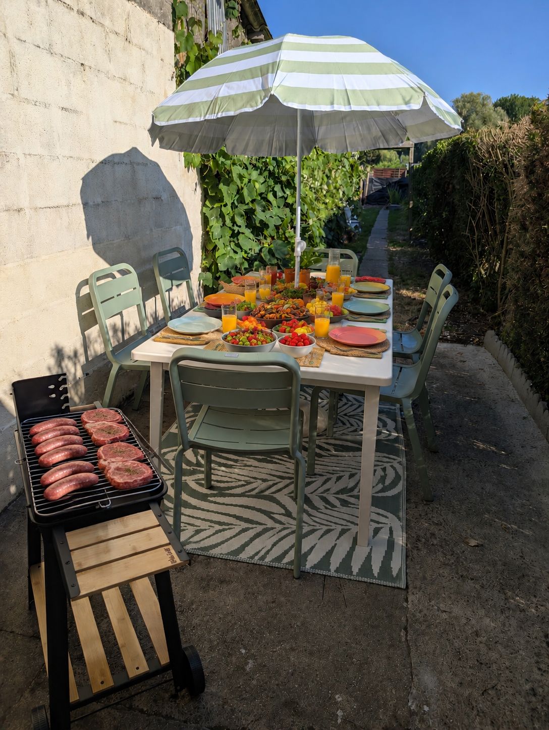 An outdoor dining area with a table set for a meal, surrounded by chairs, and a grill with sausages.