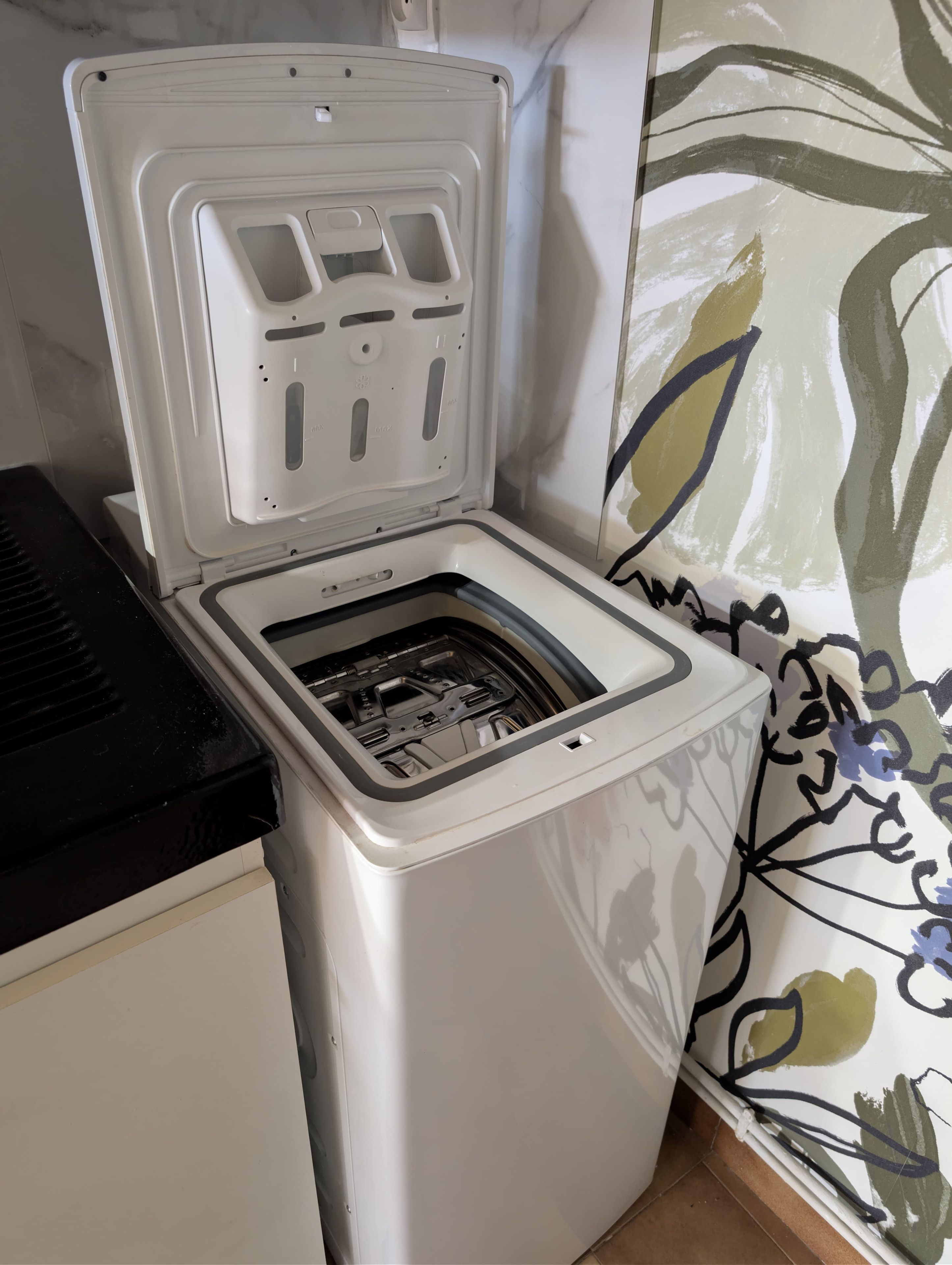 Une machine à laver compacte, de couleur blanche, dotée d’une porte transparente, située dans une pièce dont le mur est décoré d’un motif.