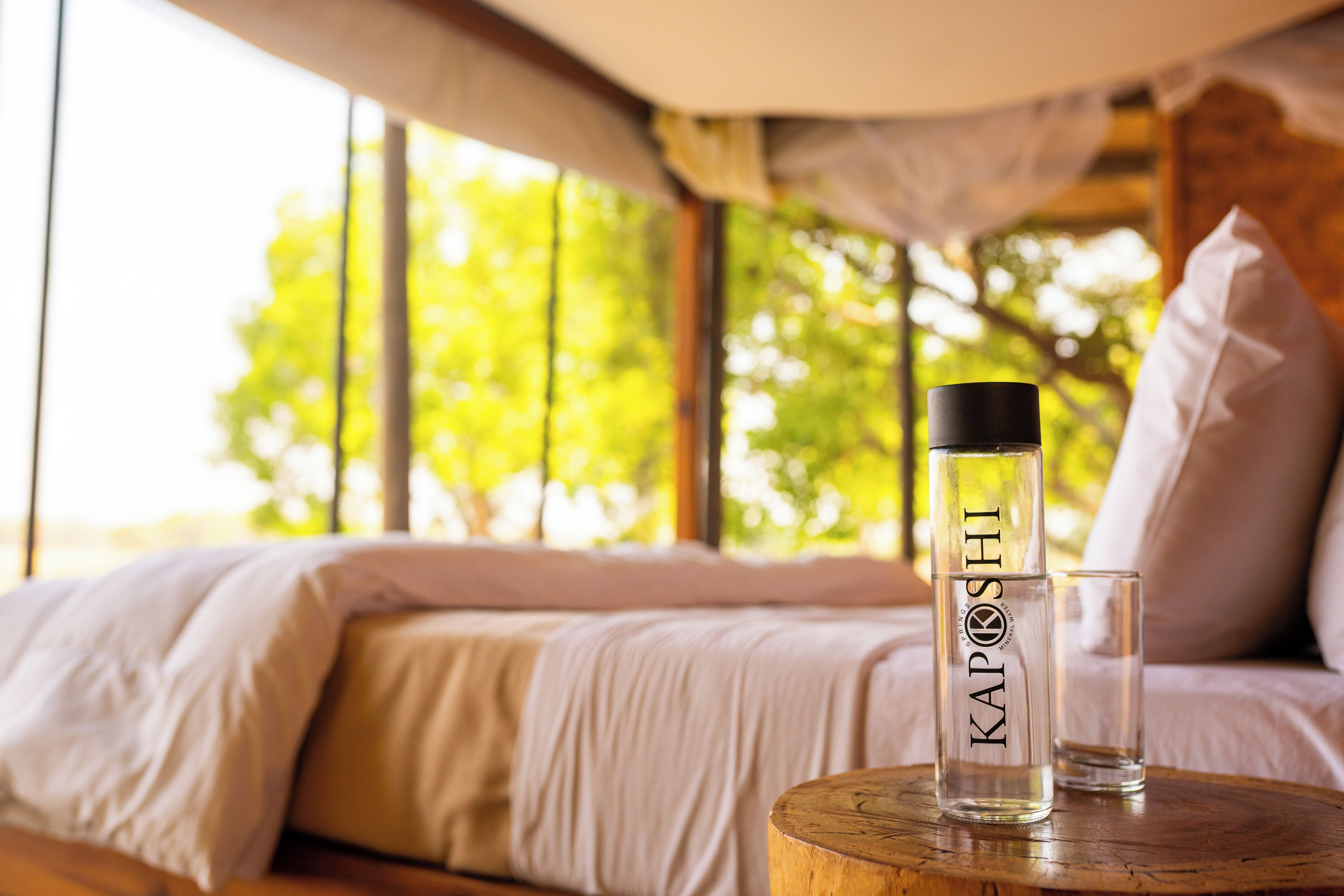 A bed with a white comforter and pillows, a clear bottle with 'KAPOS' text, and a glass on a wooden table.