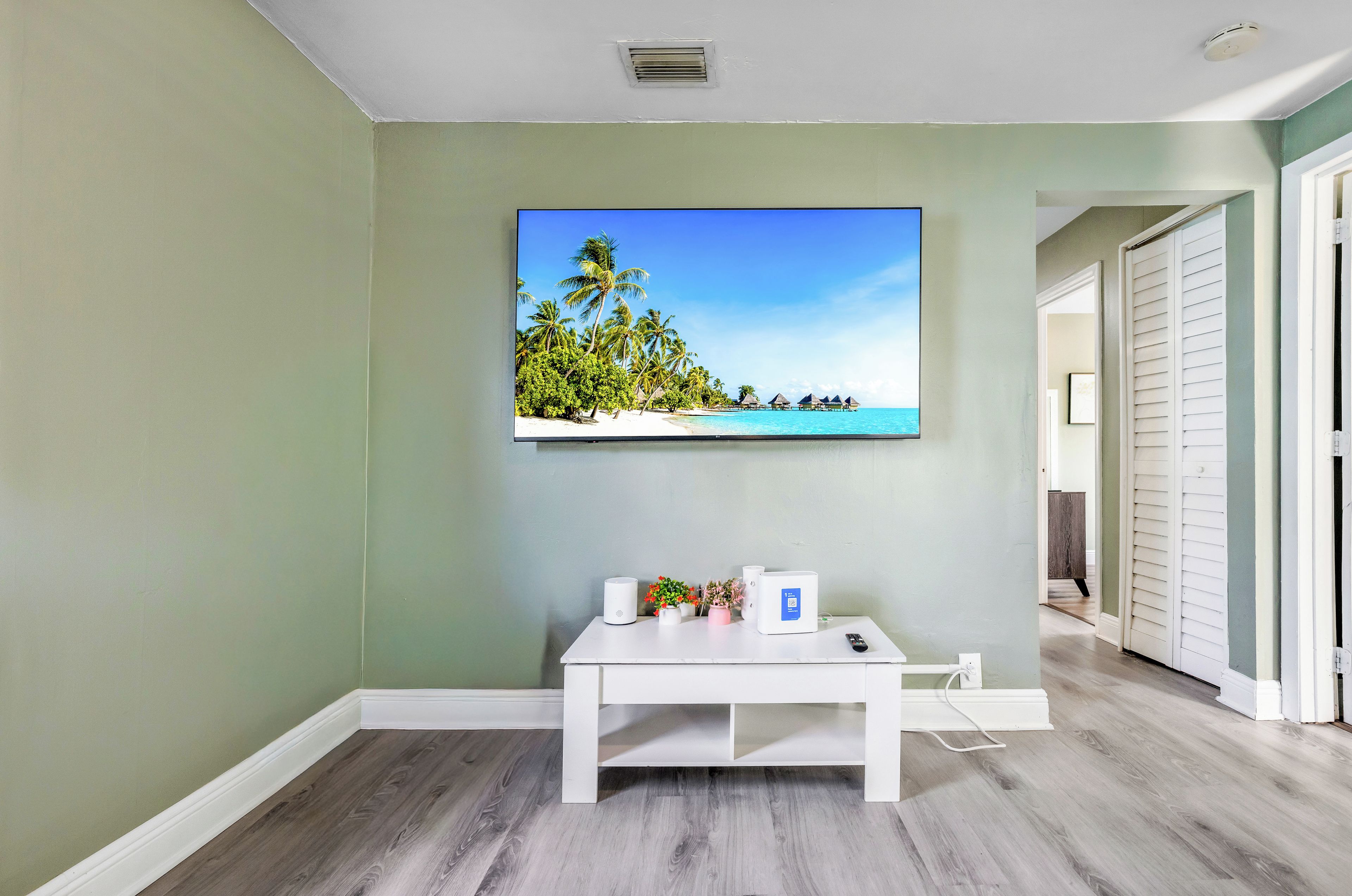 A room with a flat-screen TV mounted on the wall, a white console table with a remote control, and a door leading to another room.