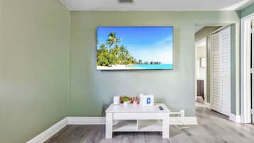 A room with a flat-screen TV mounted on the wall, a white console table with a remote control, and a door leading to another room.