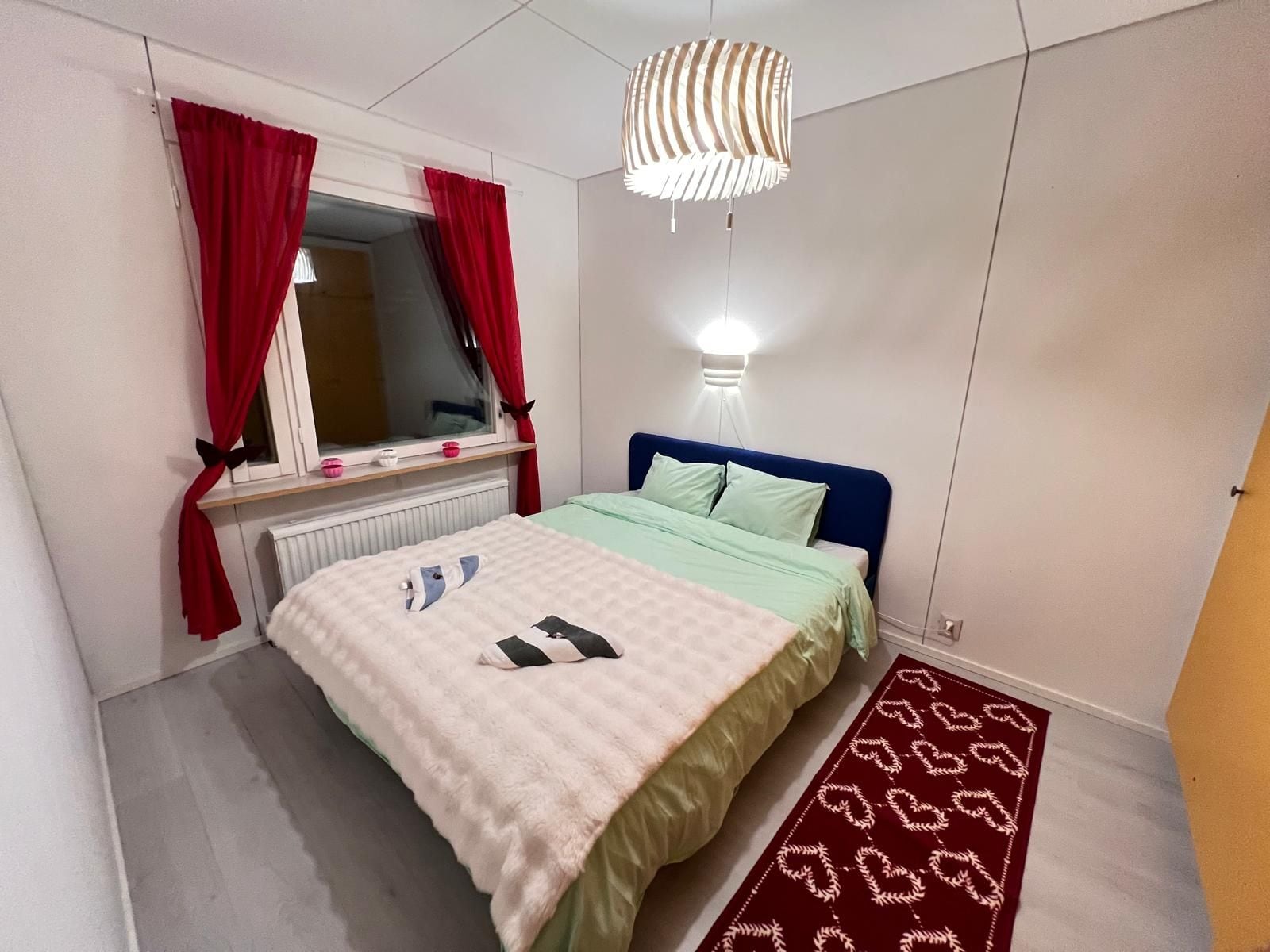 A bedroom with a bed, a window with red curtains, a bedside lamp, and a patterned rug.