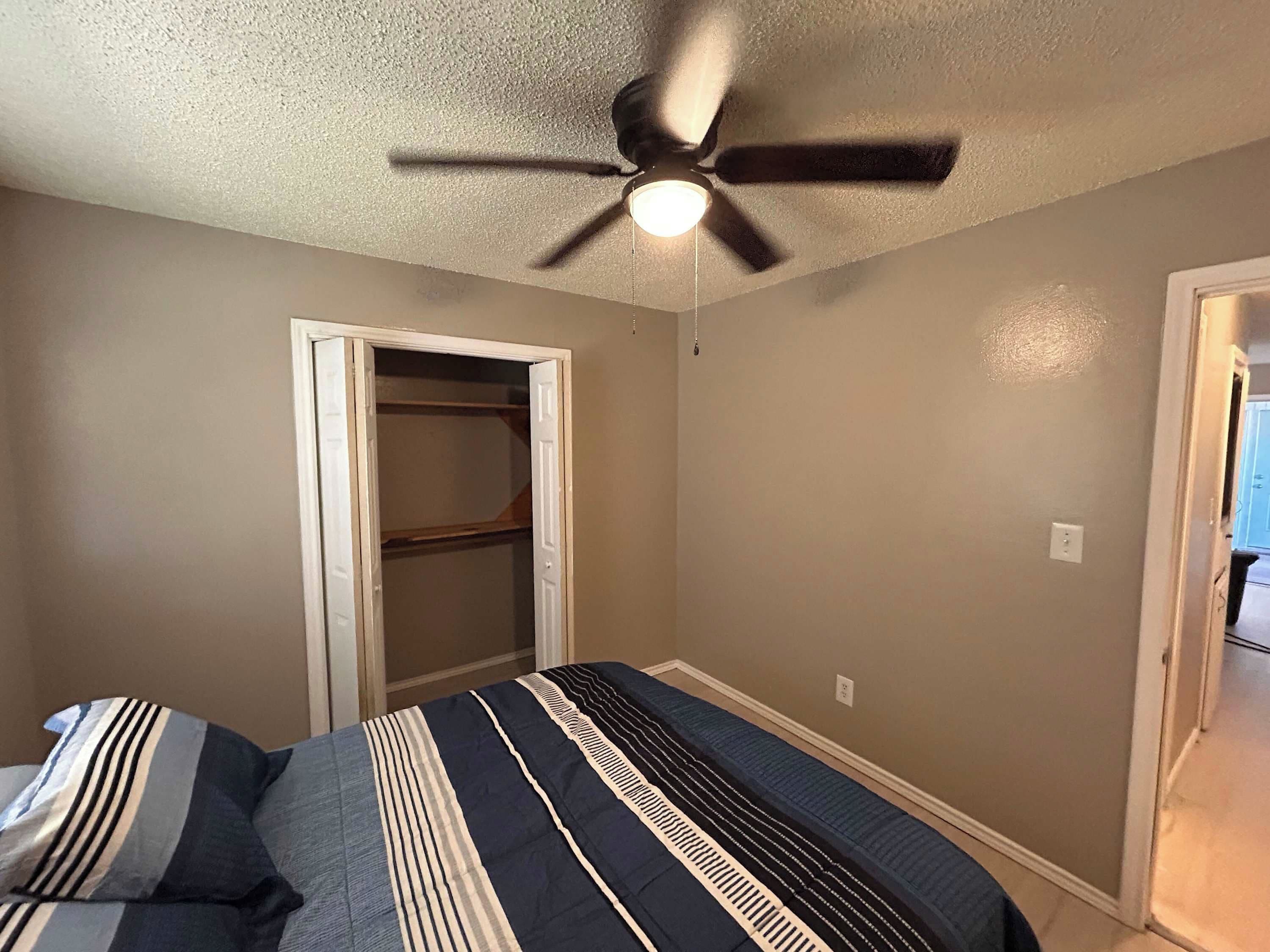 A bedroom with a bed, a ceiling fan, a closet, and a door leading to another room.
