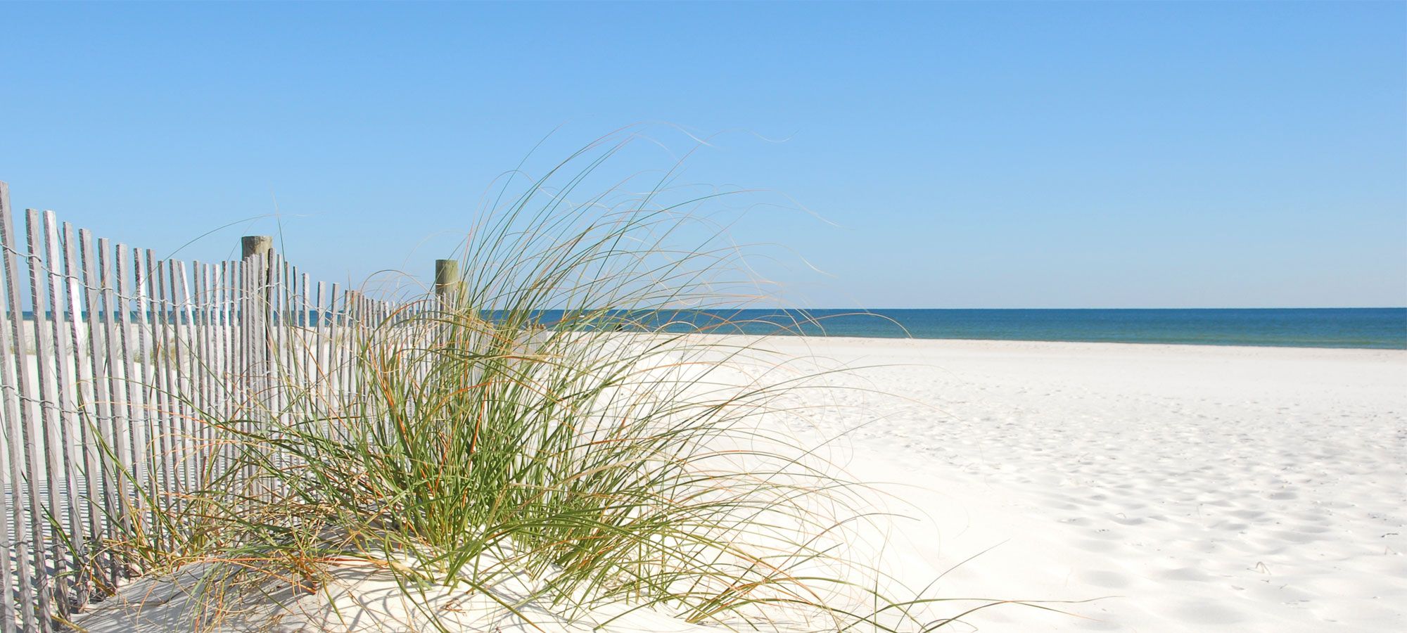 A sandy beach with a wooden fence and some grass in the foreground.