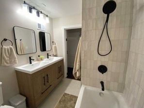 Un baño con lavamanos doble, un espejo, toallero, ducha con cabezal de ducha manual y un inodoro.