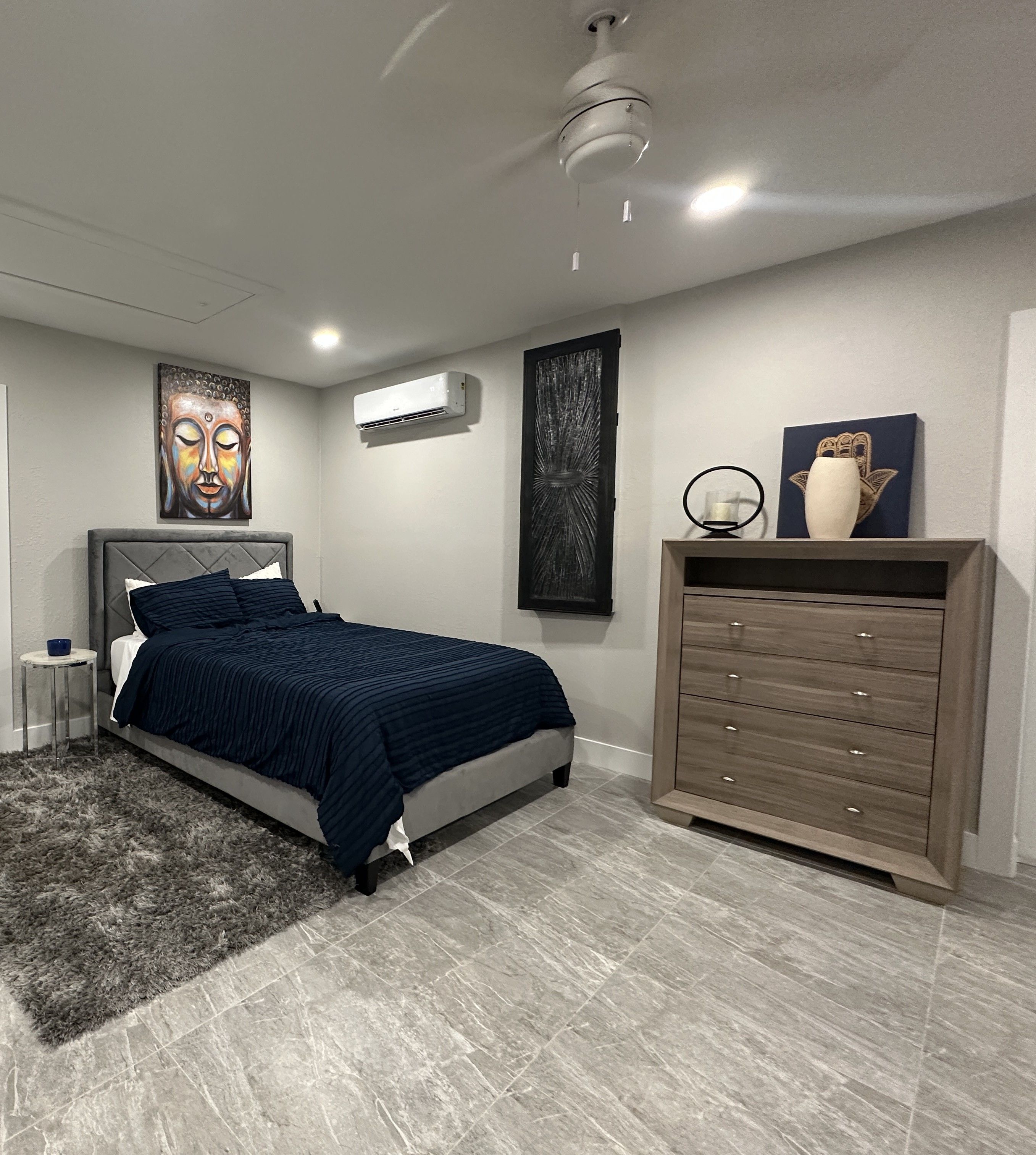 A bedroom with a bed, a dresser, a wall-mounted air conditioner, and a ceiling fan.