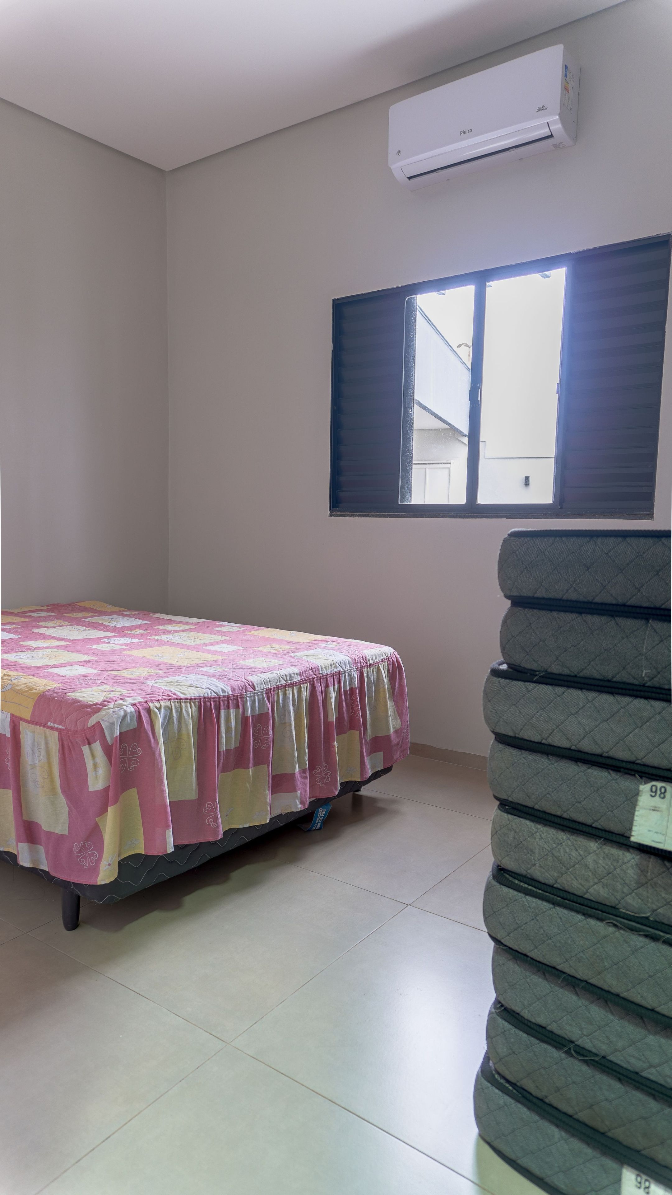 A room with a bed, an air conditioner, a window with blinds, and a stack of mattresses.