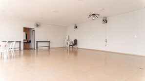 Een ruime kamer met betegelde vloer, een tafeltje met stoelen, een wandventilator en een plafondlamp.