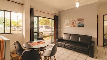 A modern living room with a black leather sofa, a round dining table with chairs, and a large window with a view of a parked car and greenery outside.