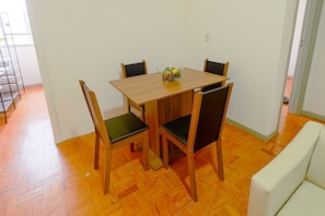 Uma sala de jantar com mesa de madeira e quatro cadeiras, um pequeno vaso com flores e uma parede branca.