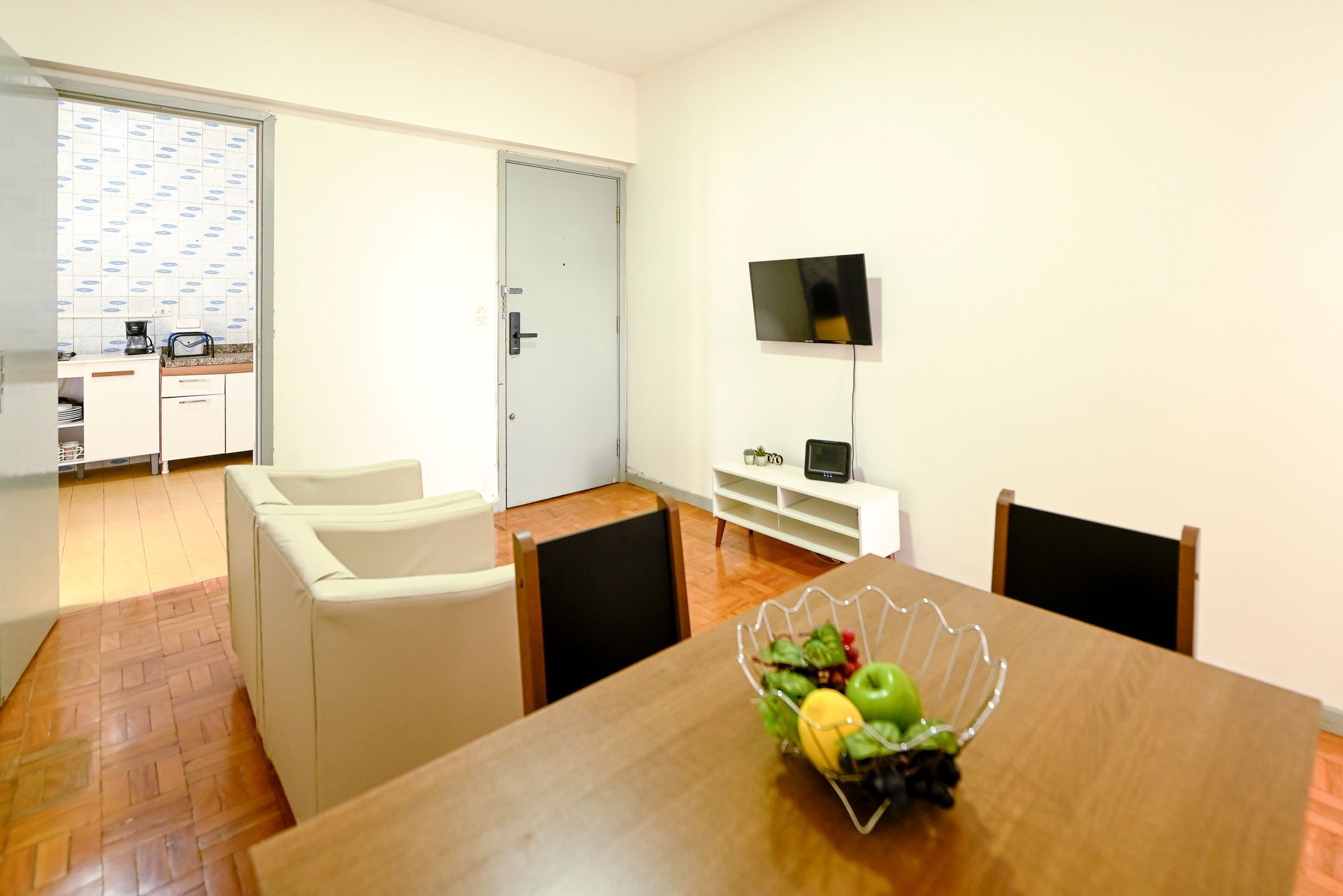 Uma área de refeições com uma mesa de madeira, duas cadeiras, um armário branco e uma TV na parede.