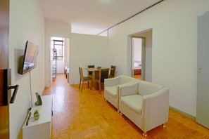 Uma sala de estar moderna com um sofá, uma mesa de jantar com cadeiras, uma televisão na parede e piso de madeira.