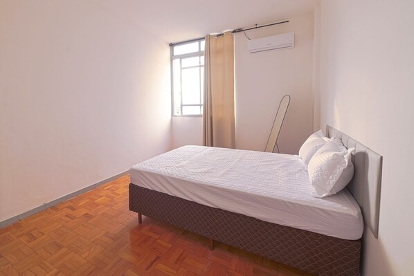 A single bed with a headboard, a window with curtains, and an air conditioning unit on the wall. - Apt with A/C near the Airport | Downtown (Campo Grande)