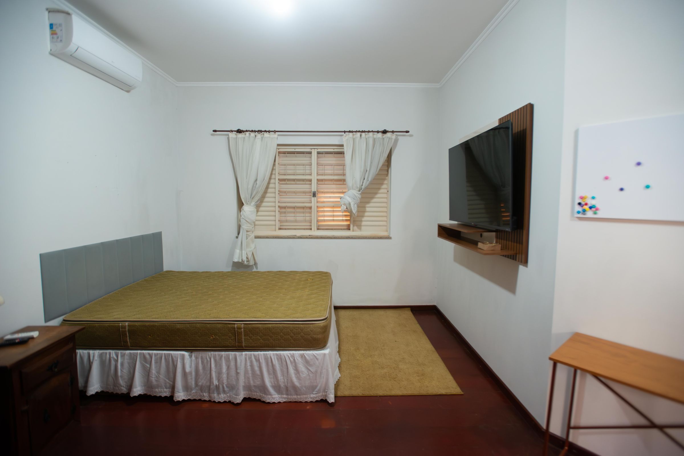 Une petite chambre avec un lit simple, une télévision, une fenêtre avec des rideaux et un tableau blanc fixé au mur.