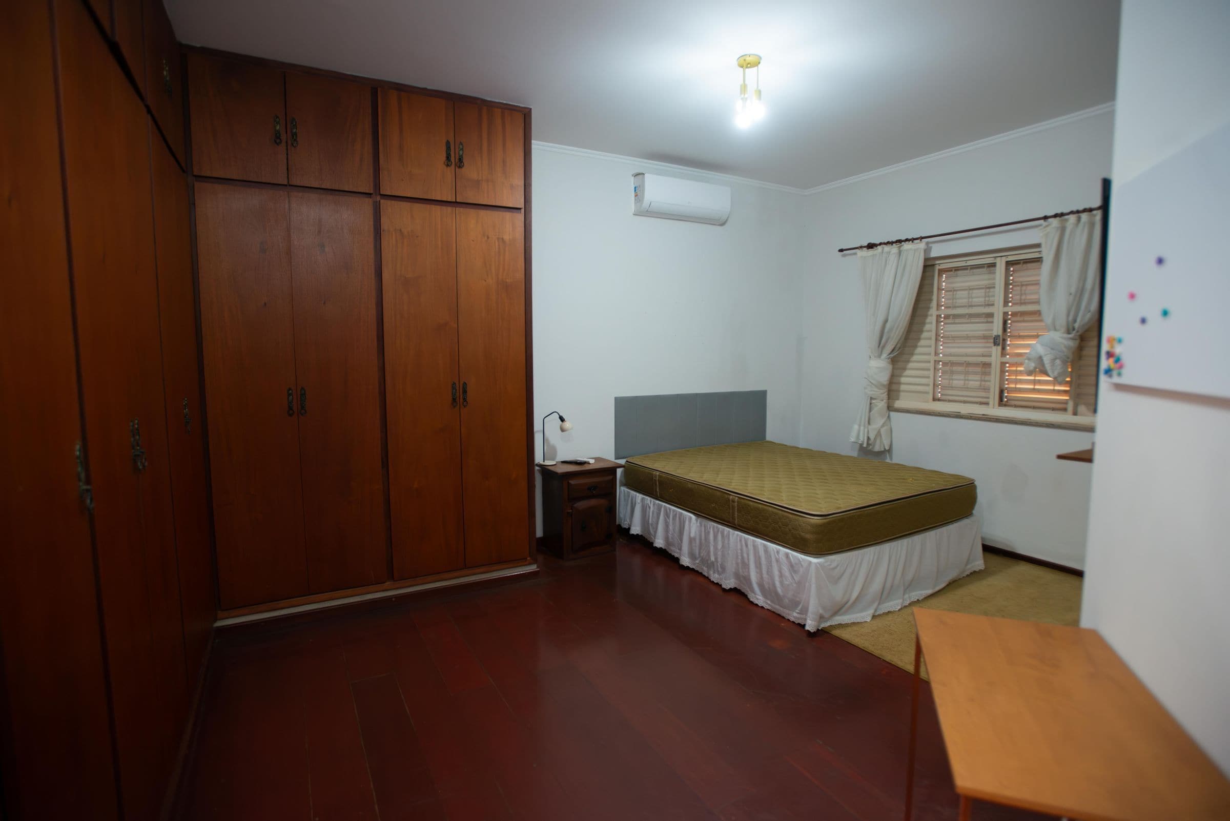 Ein Schlafzimmer mit Holzschrank, einem Einzelbett mit Matratze, einem Nachttisch, einem Fenster mit Vorhängen und einer Deckenleuchte.