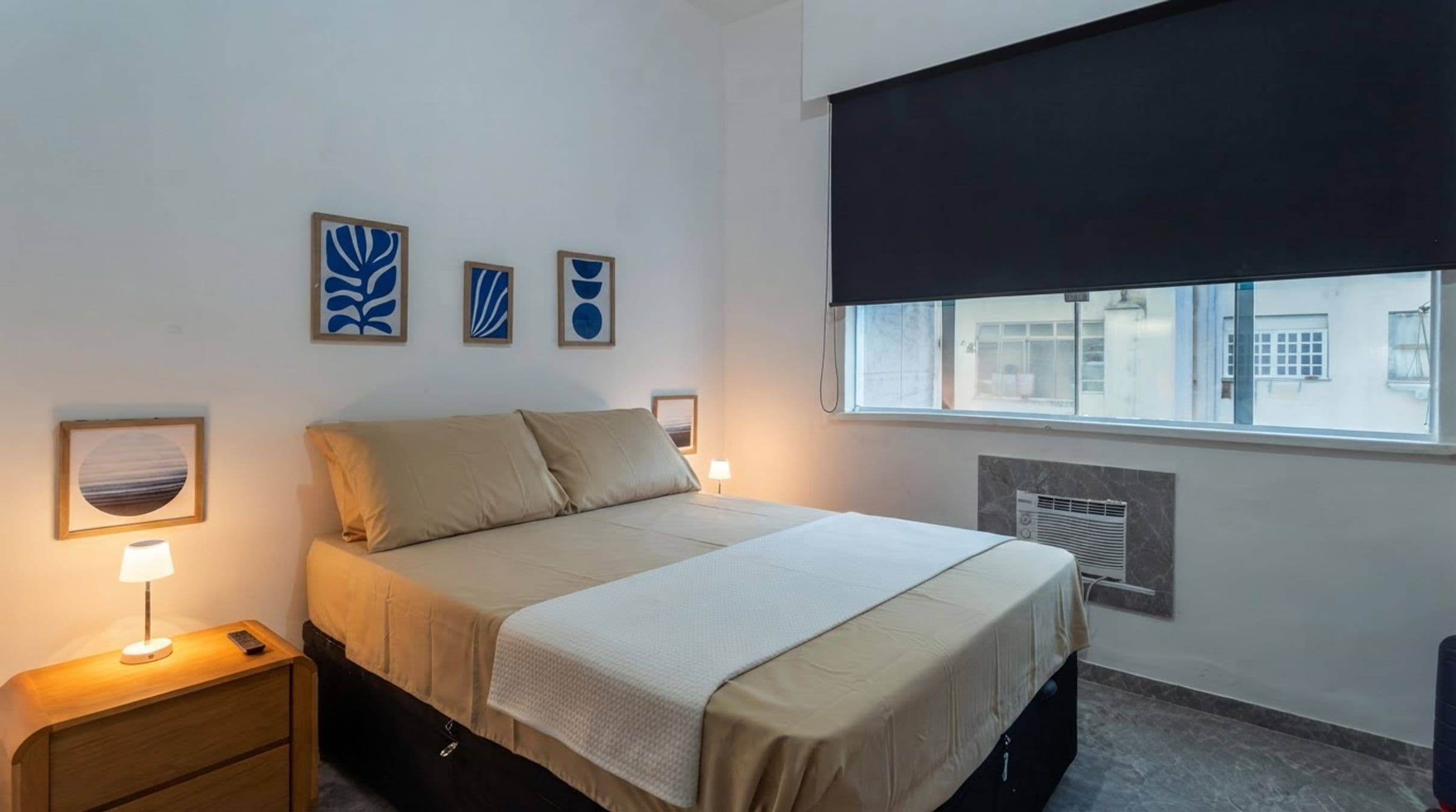 A bedroom with a bed, bedside tables, wall art, a window with a blind, and an air conditioning unit.