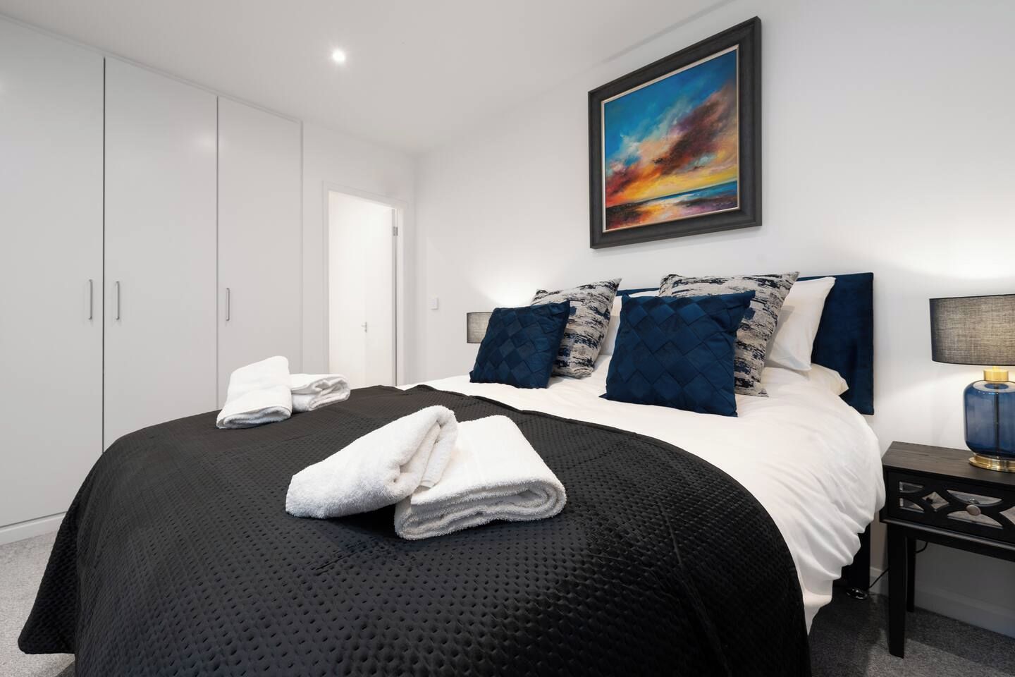 Un dormitorio con cama, almohadas, mesita de noche, lámpara y un cuadro en la pared.