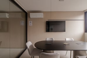 Ein Konferenzraum mit rundem Tisch, weißen Stühlen, einem an der Wand montierten Flachbildfernseher und zwei Klimaanlagen.
