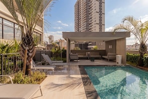 Un área de piscina en la azotea con sillones de descanso, un espacio de estar cubierto y vistas a edificios de gran altura.
