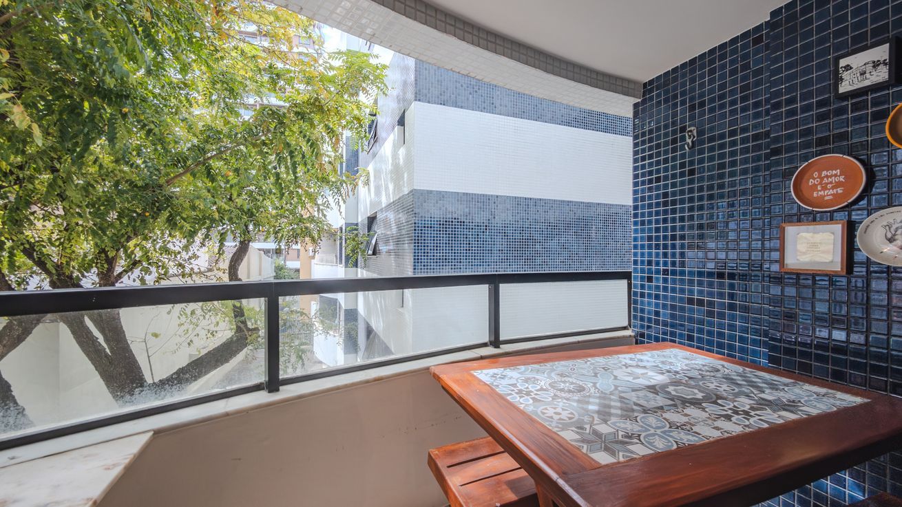 A balcony with a view of buildings and trees, a tiled wall with decorative plates, and a wooden table with a patterned tablecloth.