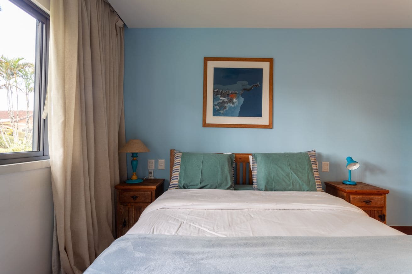 A bedroom with a bed, a nightstand with a lamp, a framed picture on the wall, and a window with curtains.