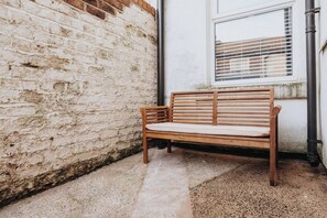 Un banco de madera frente a un muro de ladrillo blanco, con una ventana y una tubería negra.
