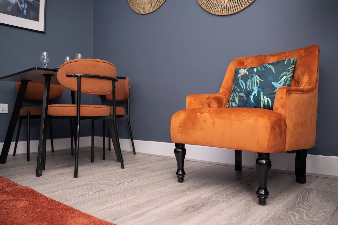Ein Zimmer mit dunkelblauer Wand, Holzboden, Tisch mit Stühlen und einem samtigen Ohrensessel mit blauem Kissen.