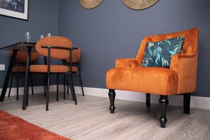 Una habitación con una pared azul oscuro, suelo de madera, una mesa con sillas y un sillón de terciopelo con un cojín azul.