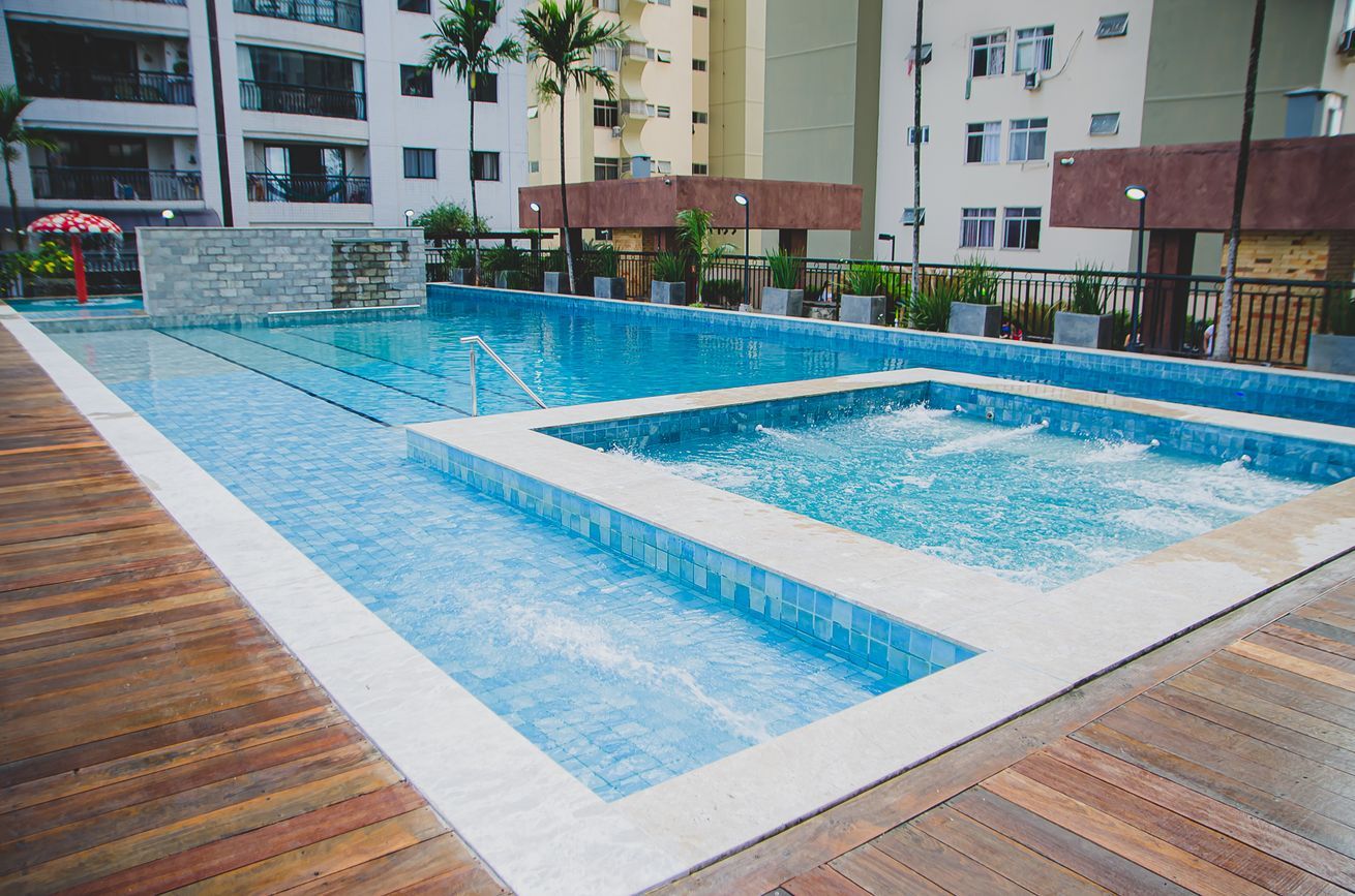 An indoor swimming pool with a wooden deck surrounding it, a stone wall, and a view of a multi-story building in the background.