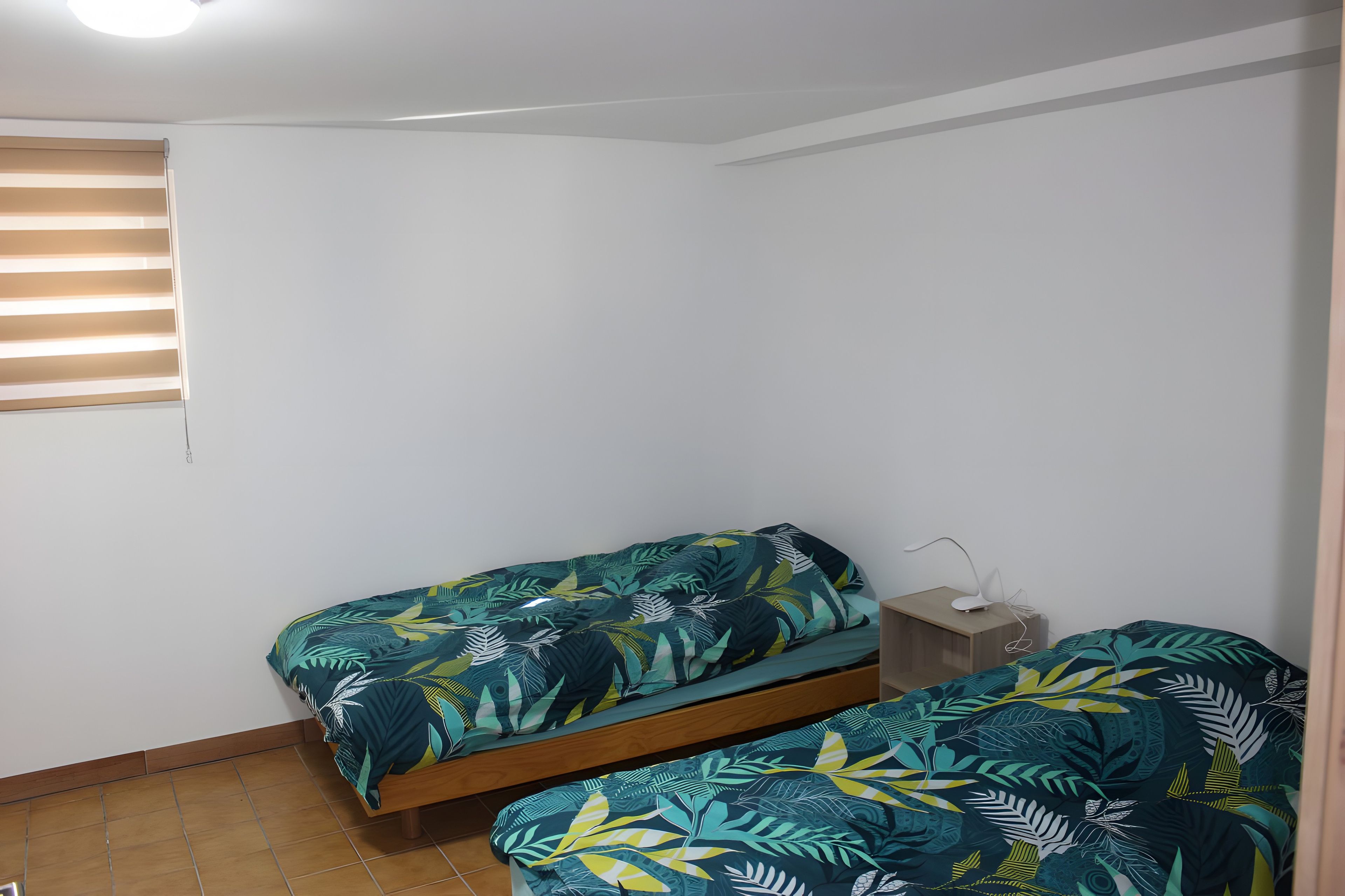 A small, simple room with two single beds, a wooden nightstand, and a white wall.