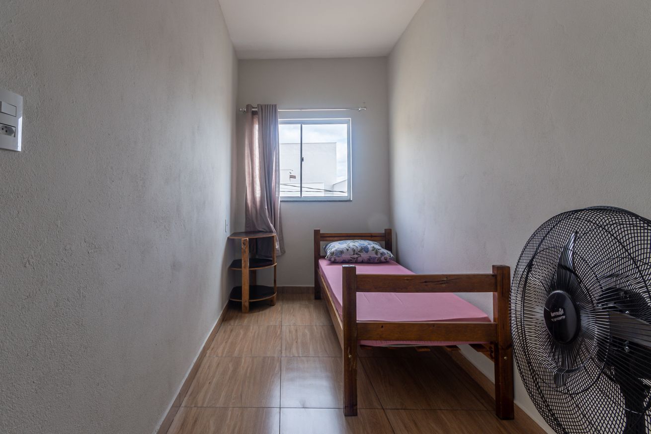 A small, narrow room with a wooden bed, a window with curtains, a standing fan, and a corner shelf.