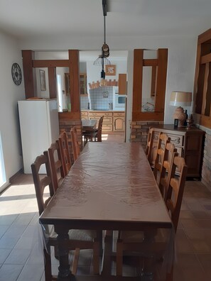 A spacious kitchen with a long dining table, wooden chairs, and a refrigerator.