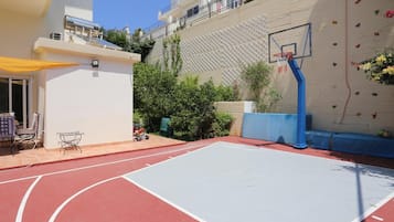 An outdoor basketball court with a climbing wall, a patio with chairs, and a building in the background.
