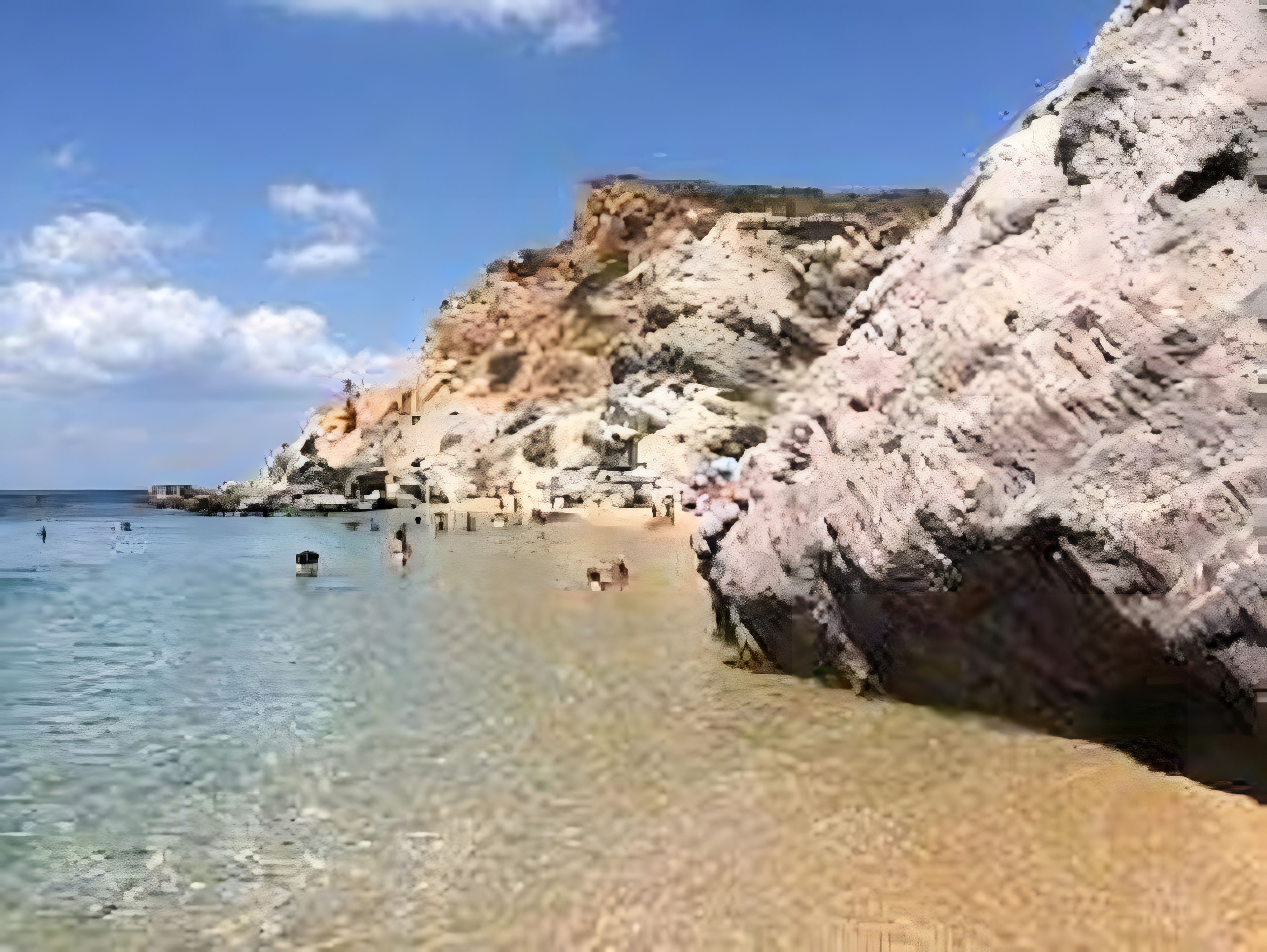 A coastal beach with clear water, rocky cliffs, and a few people swimming.