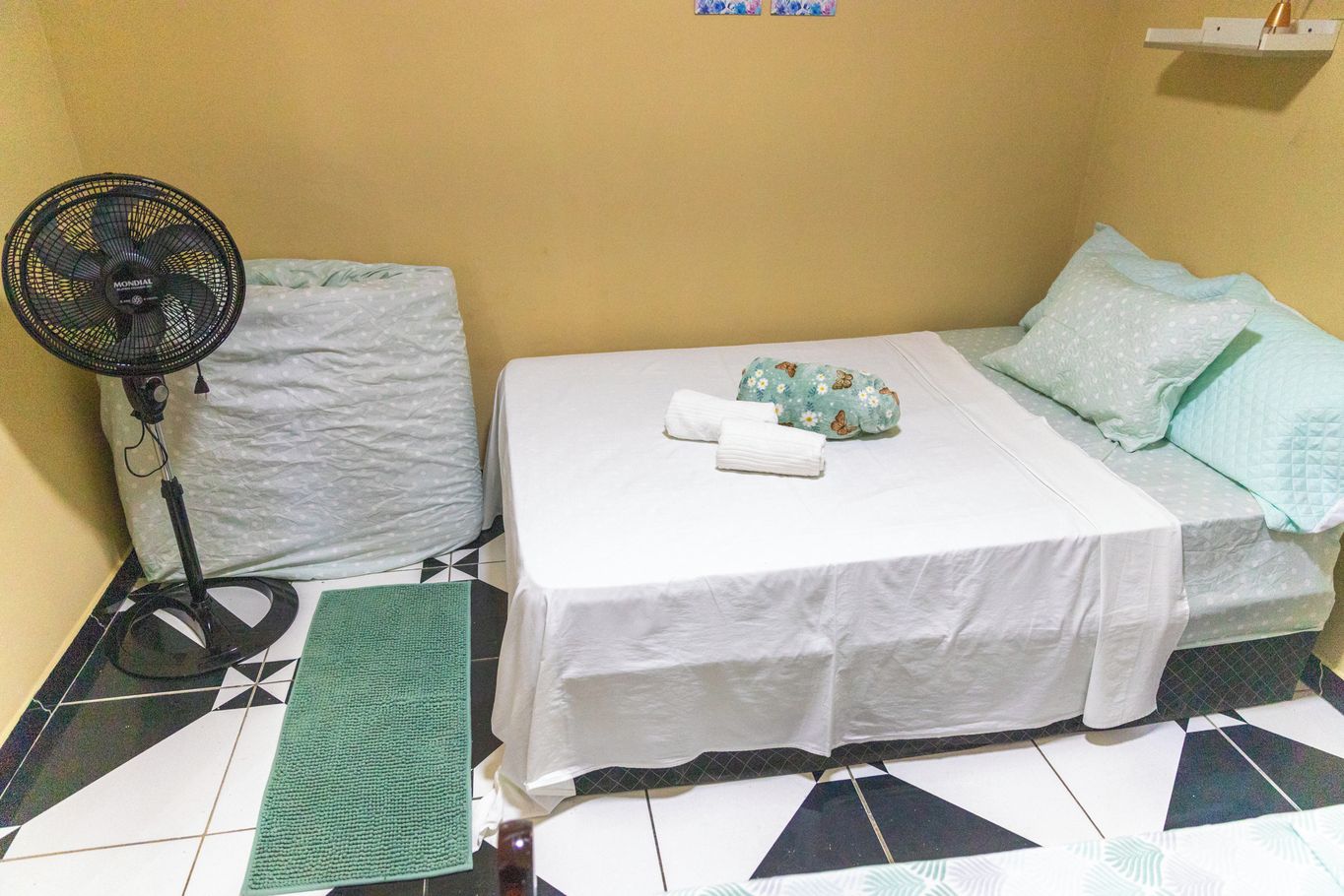 Une petite chambre d’hôtel, composée d’une seule pièce, avec un lit, un ventilateur sur pied, un tapis vert et un mur blanc.