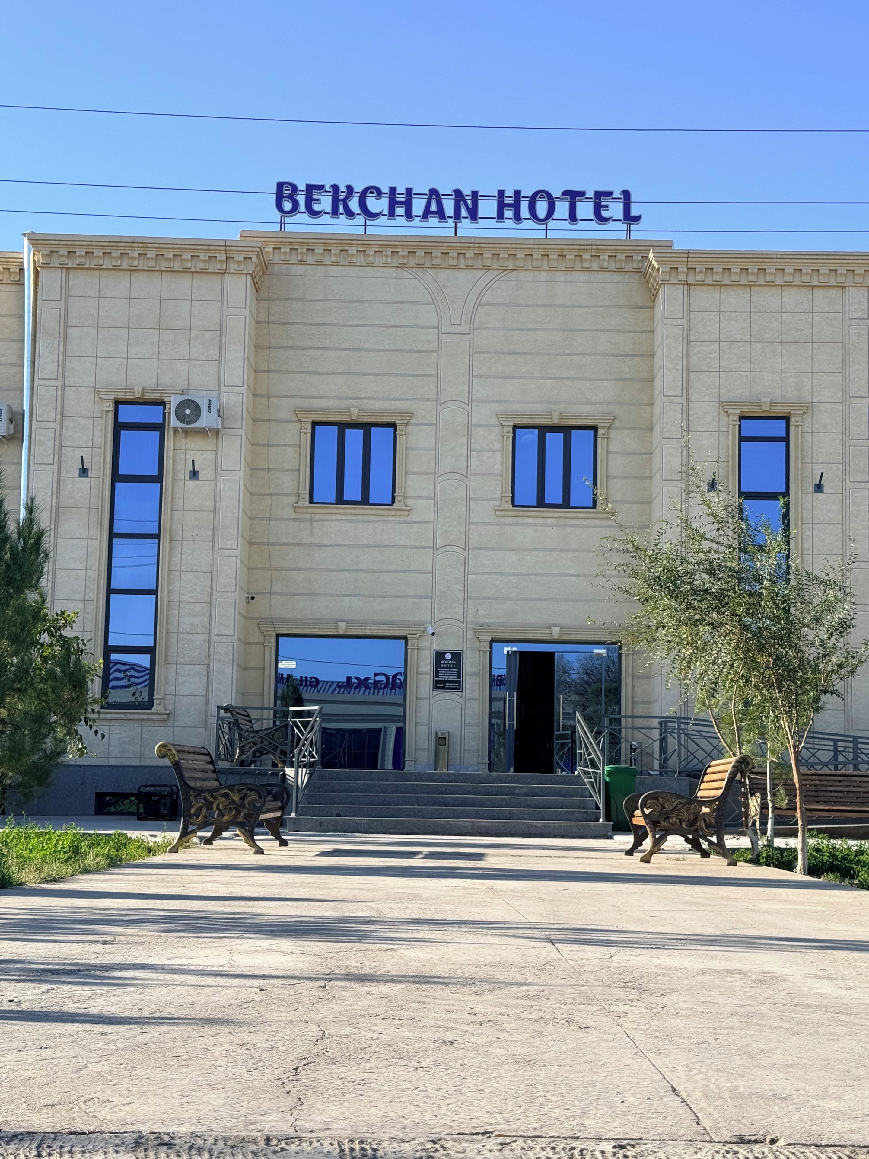Un bâtiment sur lequel on peut lire, sur une enseigne au sommet, les mots « HÔTEL BERCHAN ». Des marches mènent à l’entrée, et deux bancs sont placés devant.