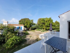 Une terrasse avec des sièges extérieurs, un bâtiment blanc avec des volets bleus et un sentier de terre menant à un grand arbre.