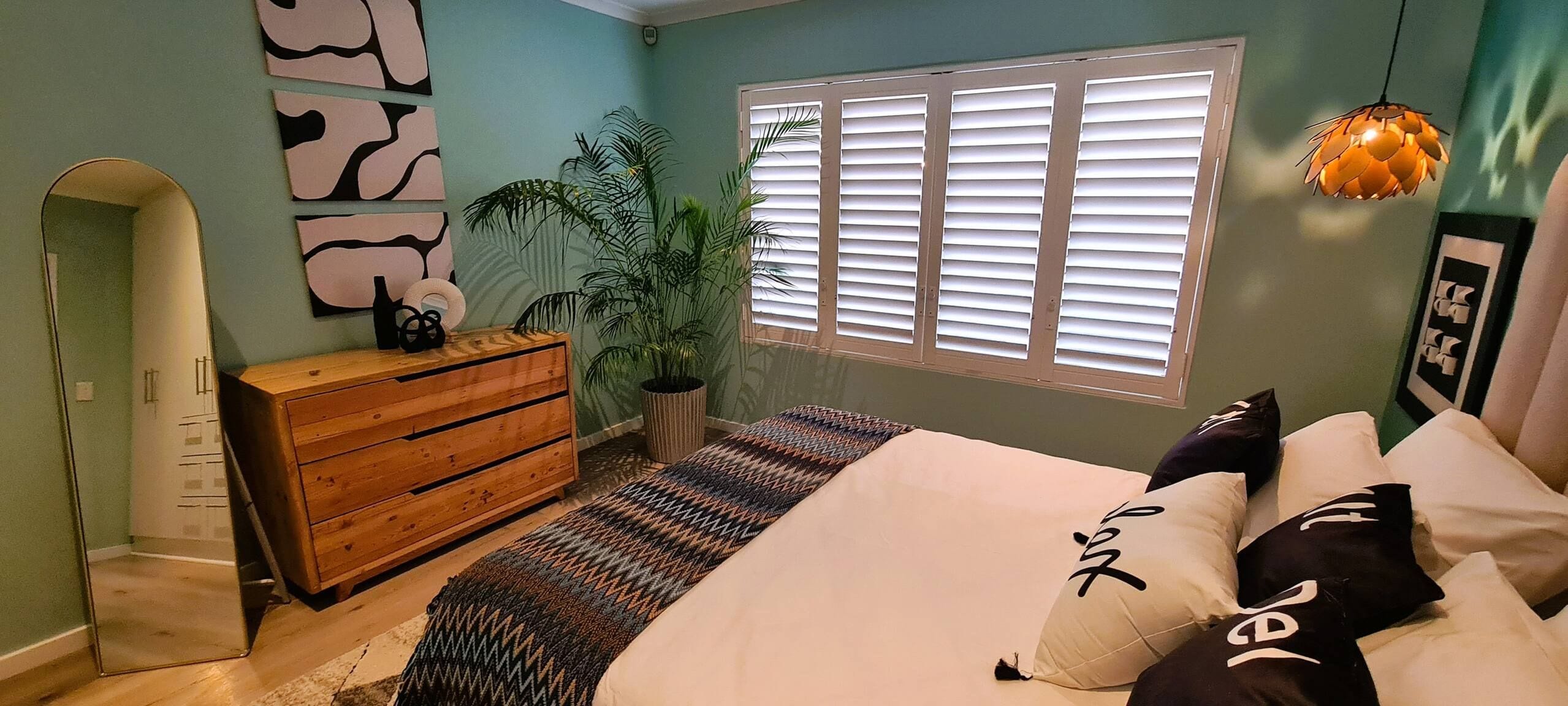 A bedroom with a bed, dresser, potted plant, and window with blinds.