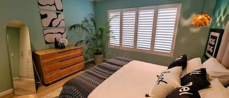 A bedroom with a bed, dresser, potted plant, and window with blinds.