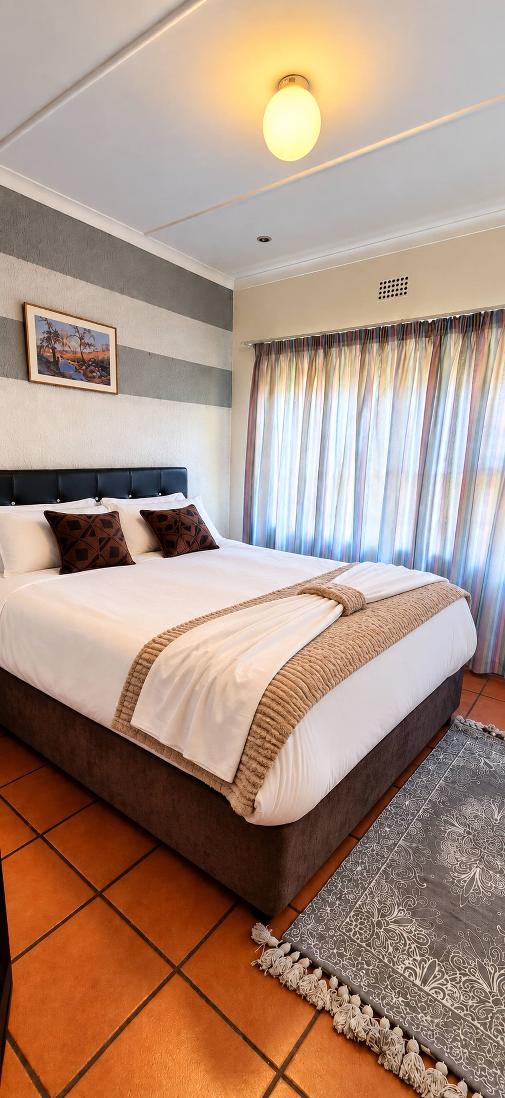 Una habitación de hotel con una cama grande, cabecero, un cuadro en la pared, una ventana con cortinas y una alfombra estampada.