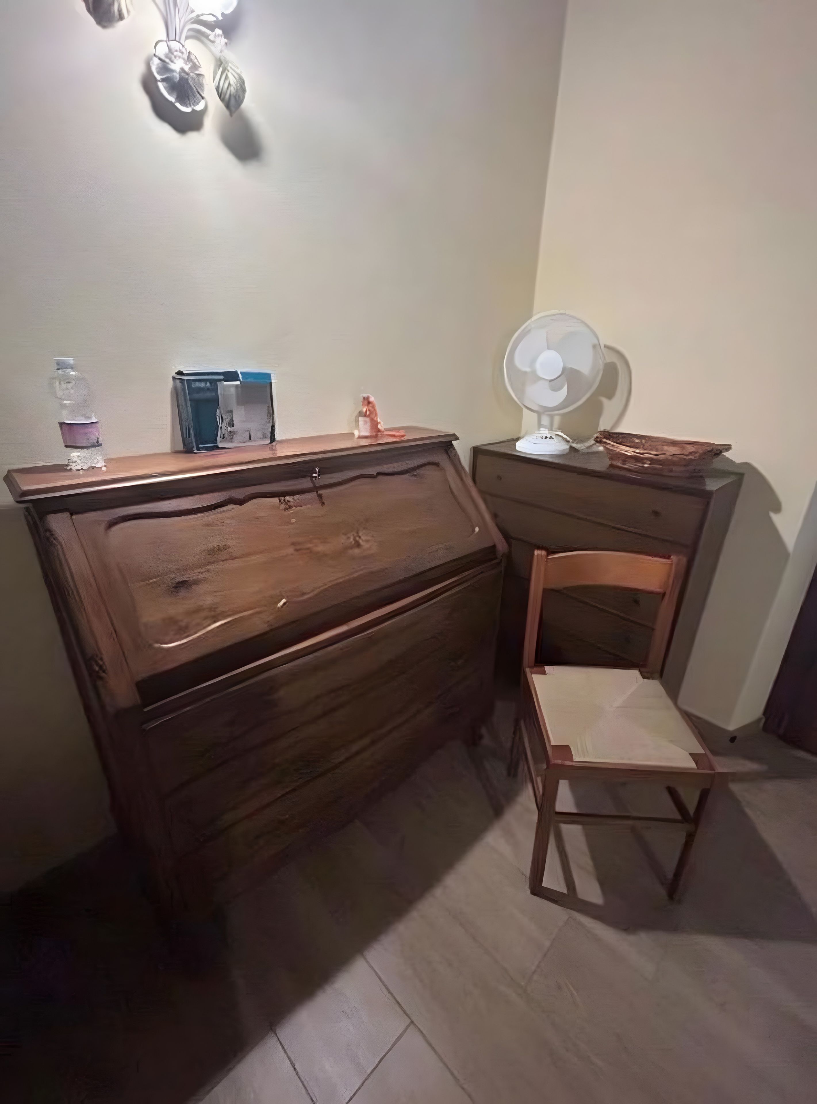 Un bureau en bois, une chaise, une lampe de bureau et un ventilateur fixé au mur.