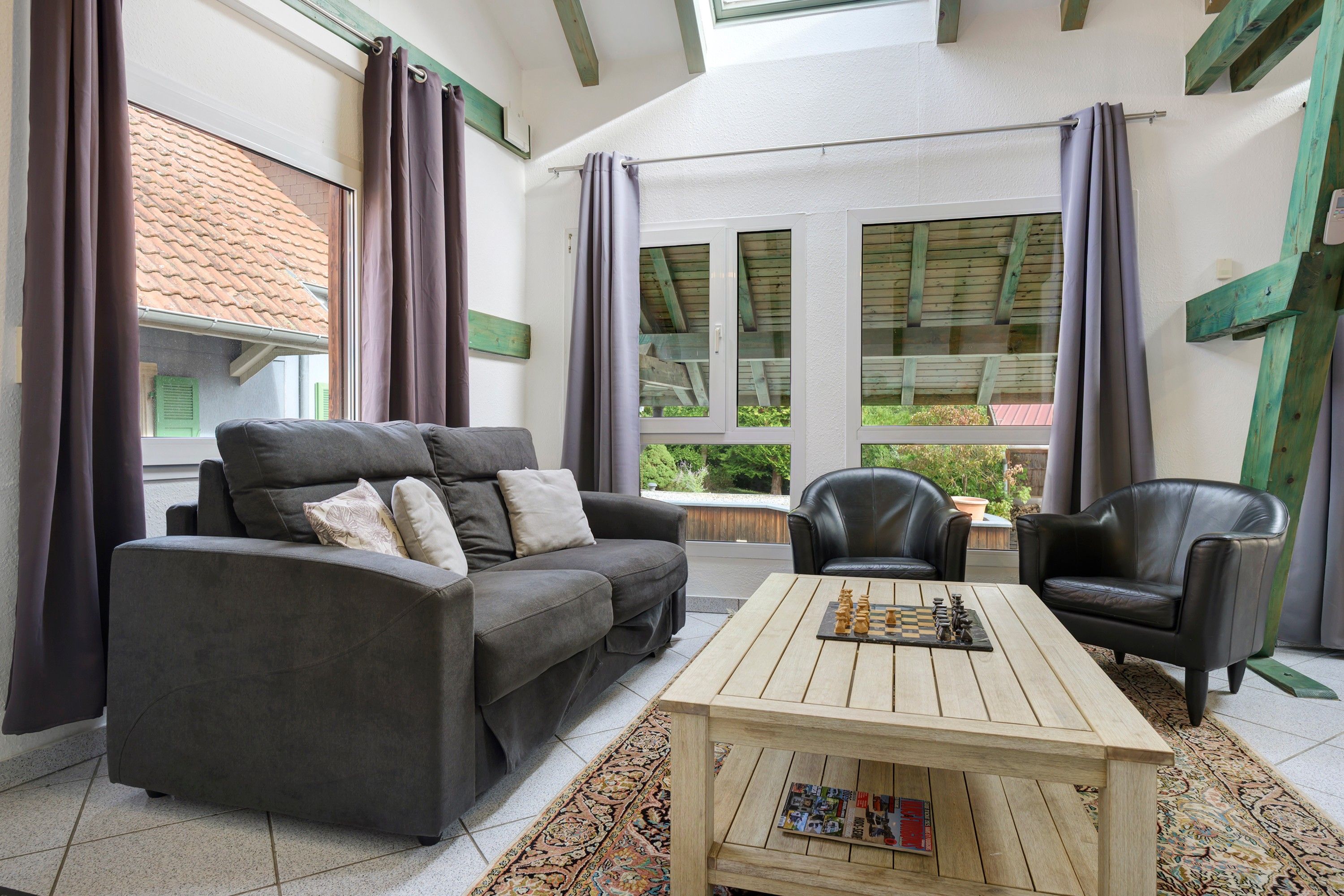 Una sala de estar acogedora con un sofá gris, dos sillones de cuero negro, una mesa de centro de madera y vistas a un jardín a través de amplios ventanales.