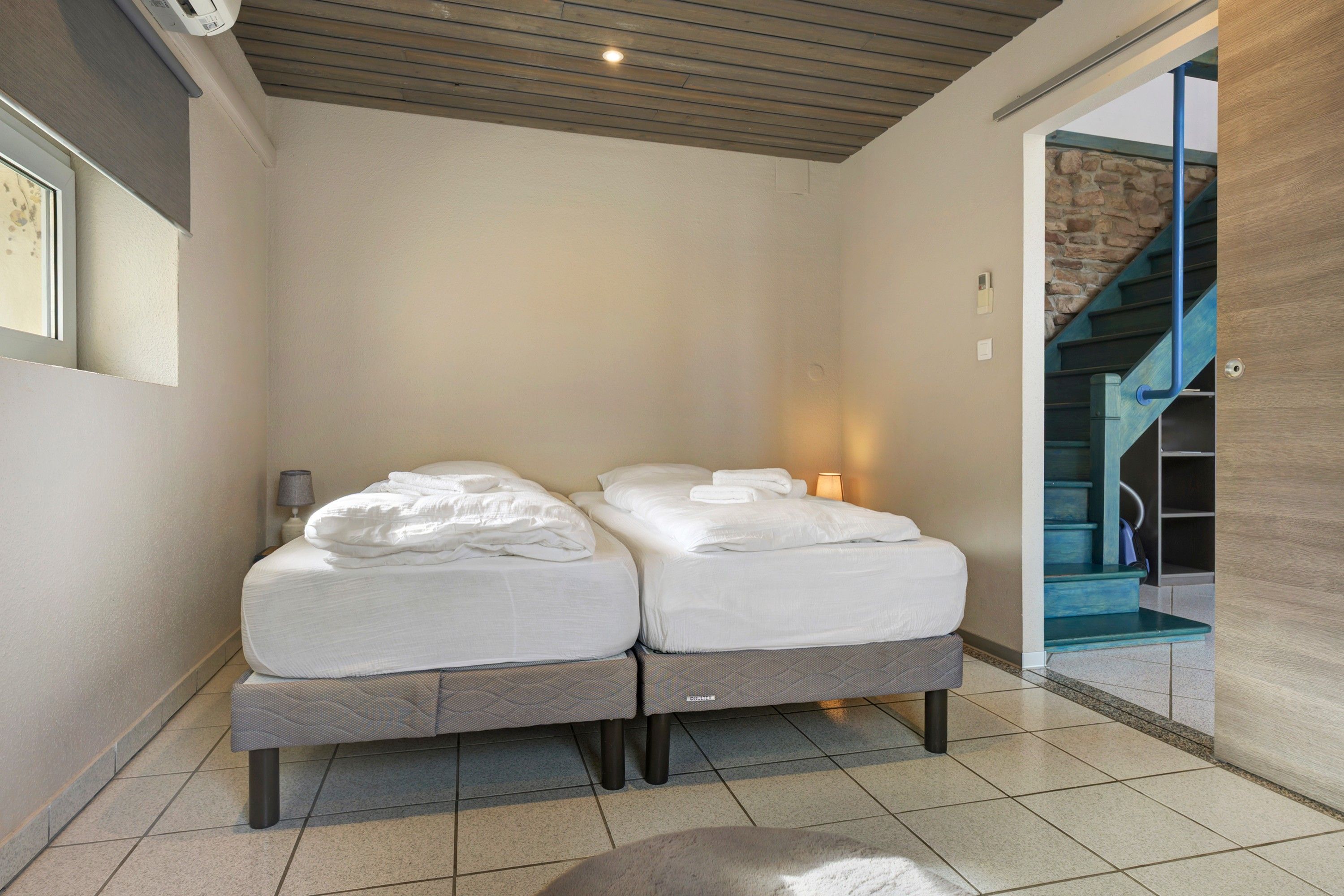 A double bed with white linens in a modern room with a tiled floor and a wooden ceiling.
