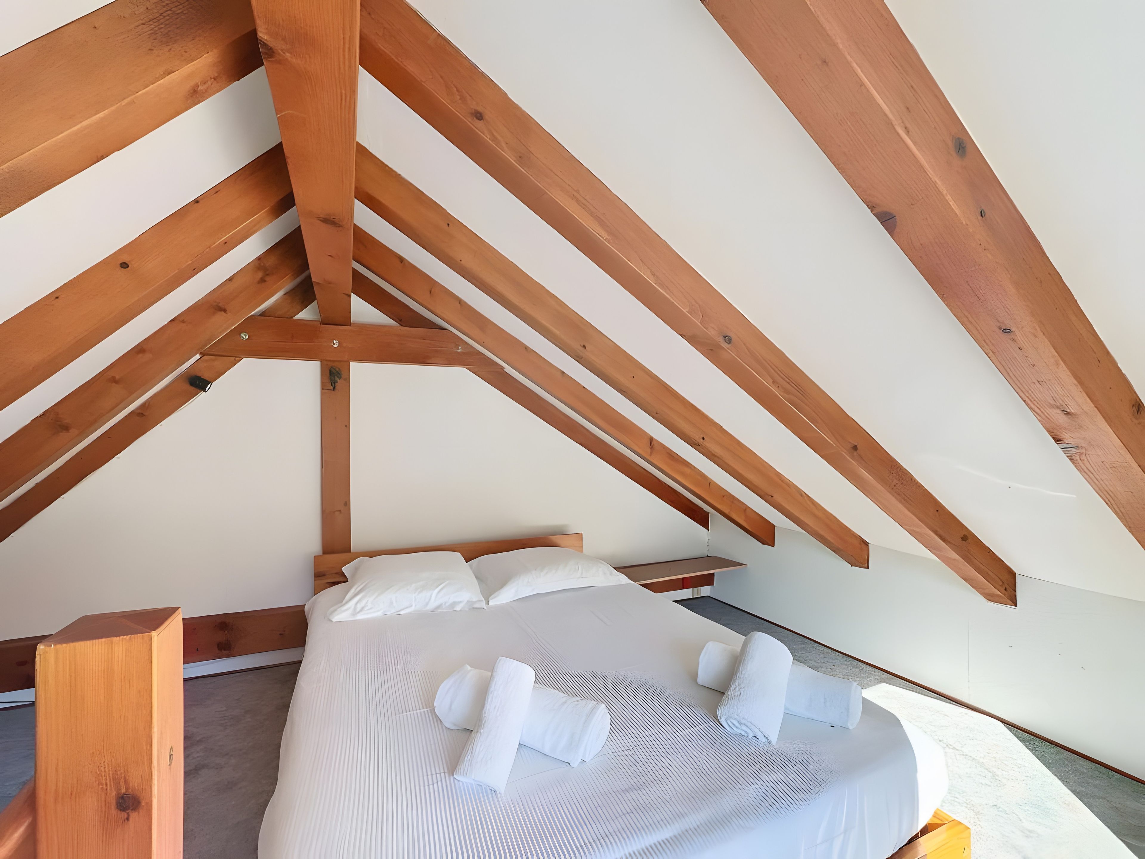 Une chambre avec un plafond en pente, des poutres en bois, un lit avec une literie blanche et une table de chevet en bois.