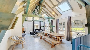 Um amplo loft com pé direito alto, vigas de madeira aparentes, uma grande mesa de jantar e vista para o exterior.