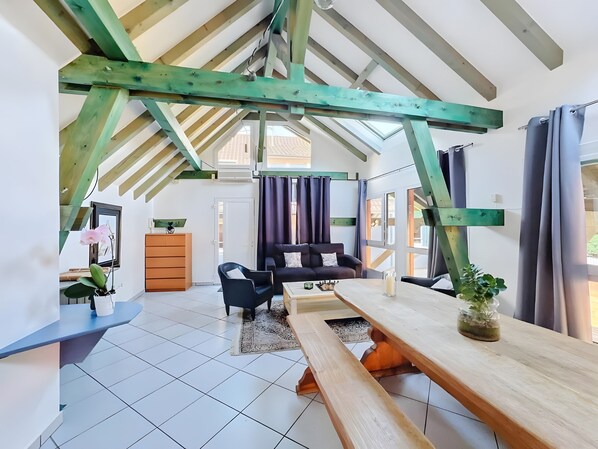 Um loft espaçoso com pé direito alto, vigas de madeira aparentes, área de refeições com mesa de madeira, sofá e uma pequena mesa lateral.