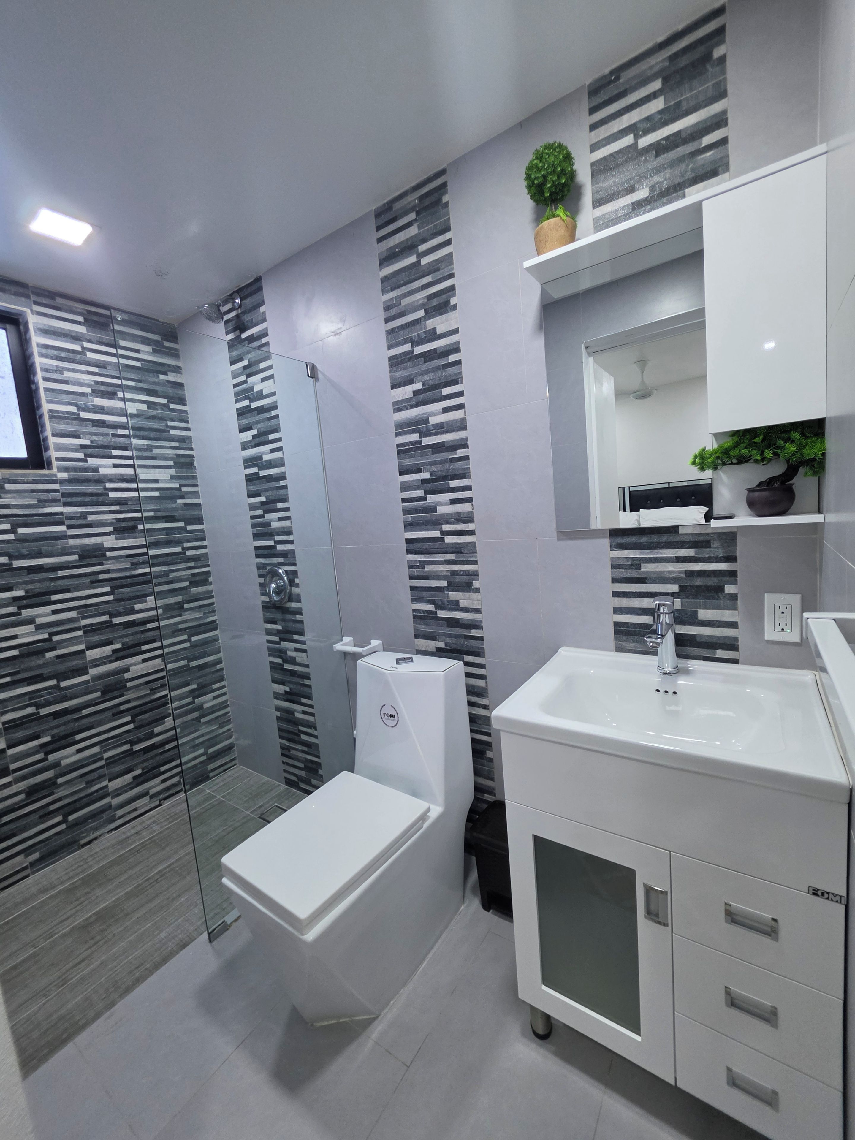 Een moderne badkamer met een glazen douchecabine, toilet, wastafel met spiegel erboven en een potplant op het aanrecht.