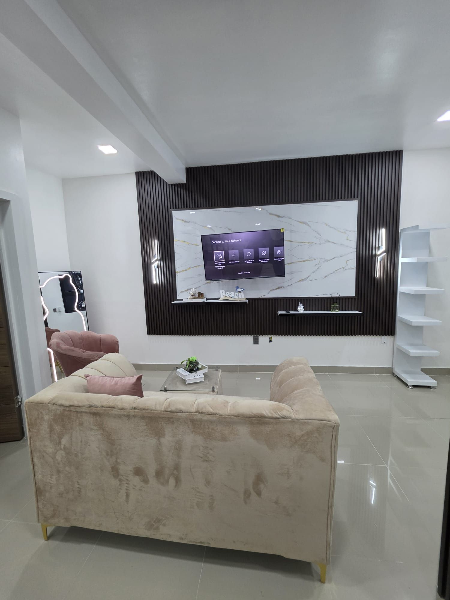 A modern living room with a large flat-screen TV mounted on the wall, a beige sofa, a coffee table with a plant, and a minimalist shelving unit.